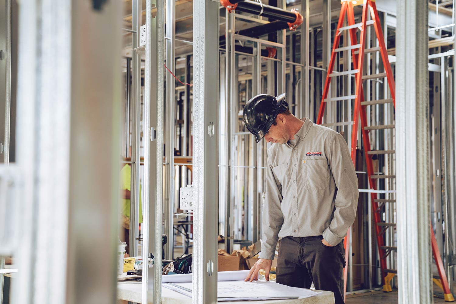 A man in a hard hat reviewing blueprints at an indoor construction site with metal studs and red ladder.