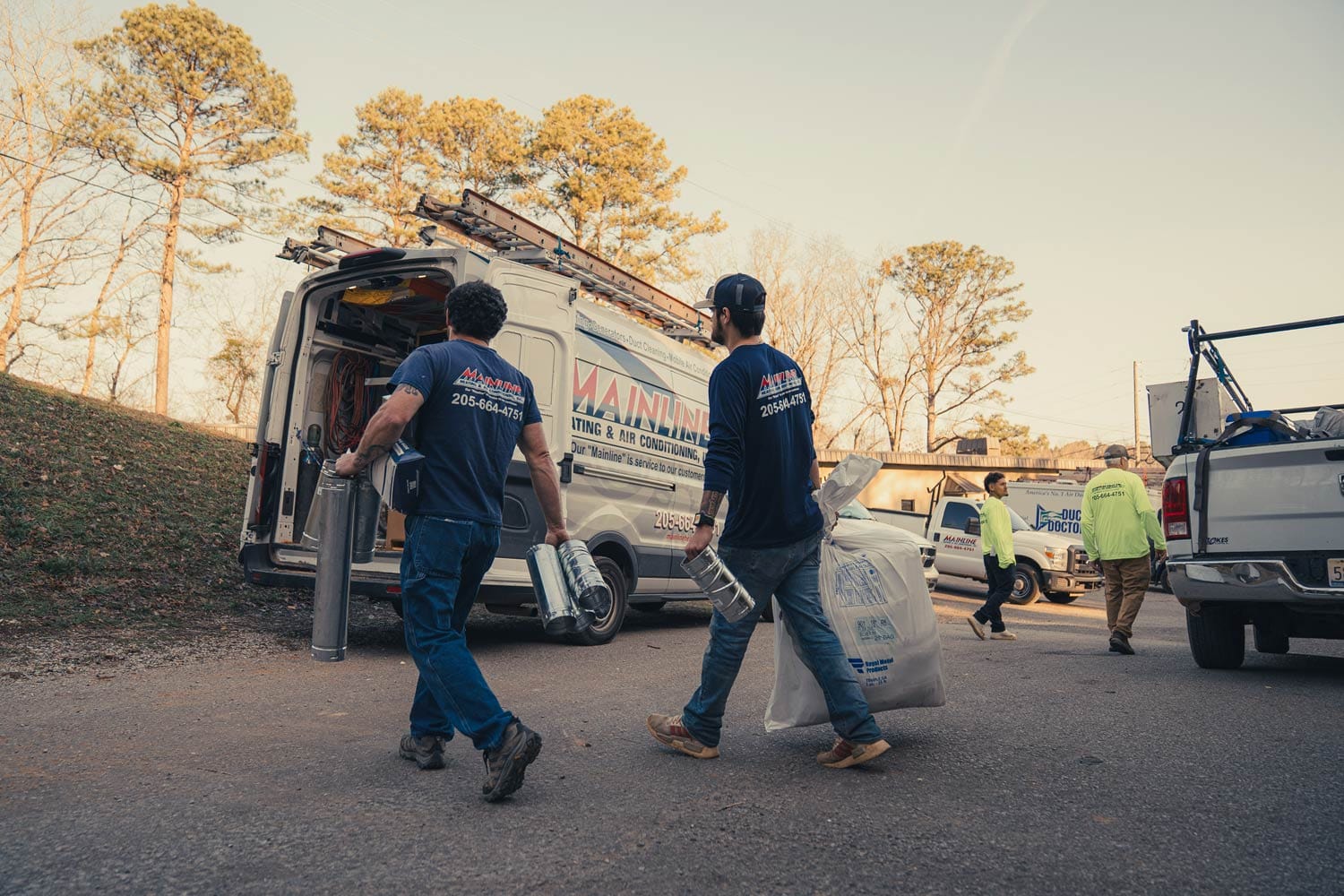 Two HVAC technicians carrying duct pipes and insulation toward a Mainline heating and air conditioning service van